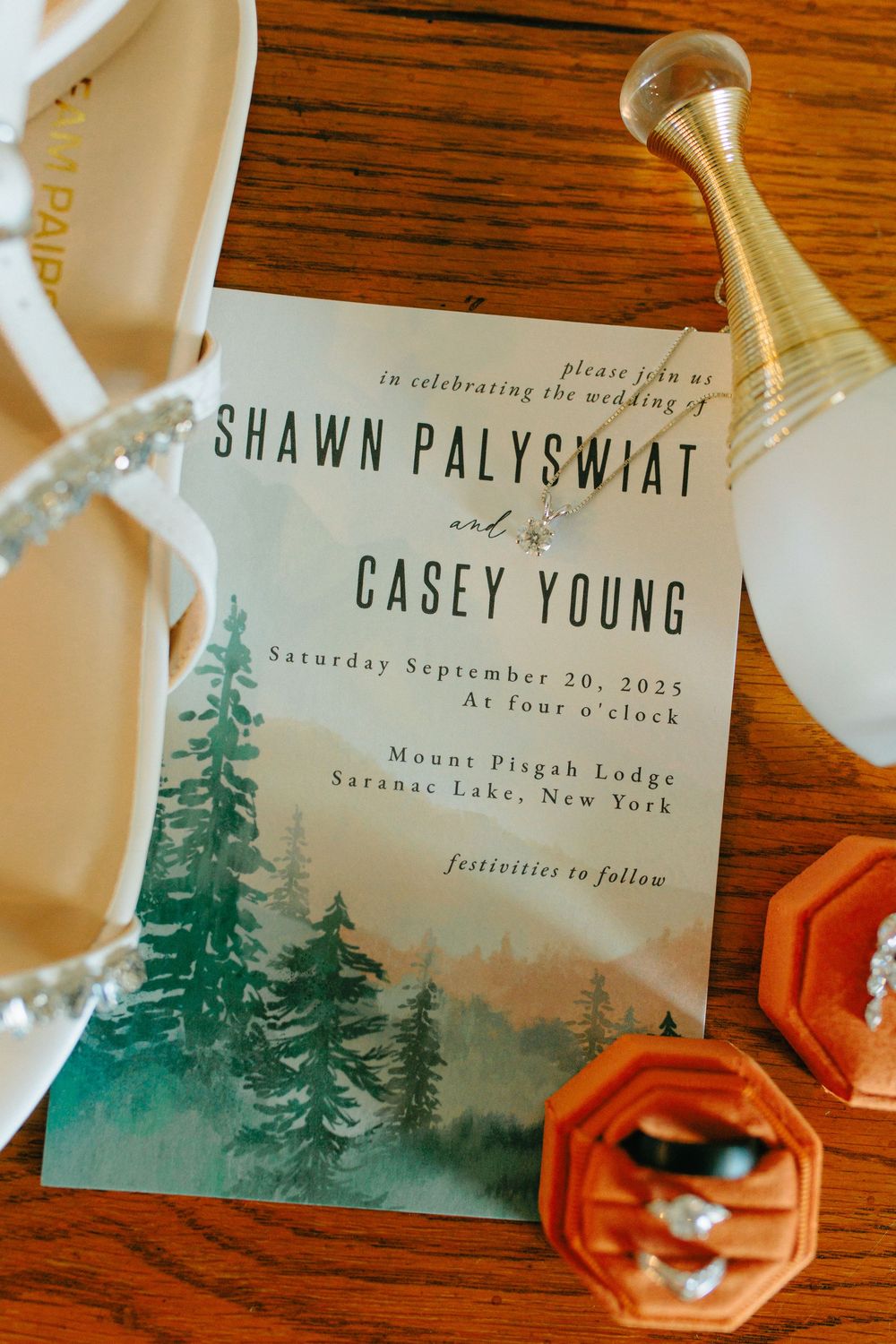 Wedding invitation featuring watercolor pine trees and Mount Pisgah Lodge details on wooden table with decor.