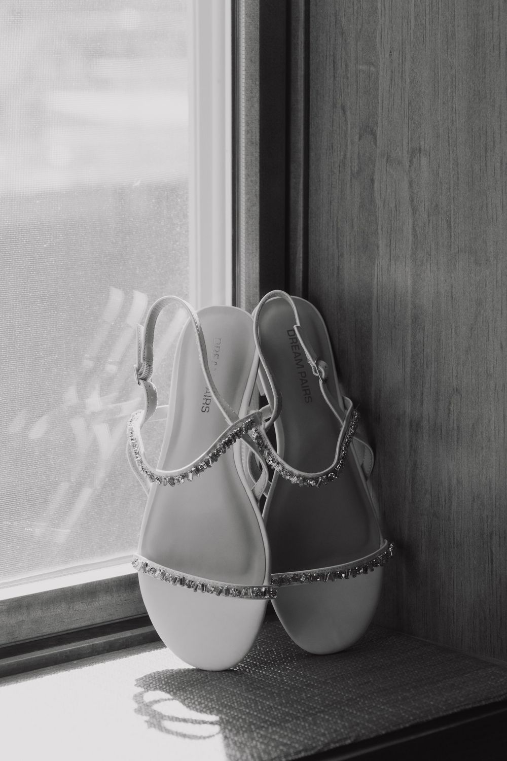 Elegant bridal sandals with crystal embellishments sit on a windowsill in black and white photography.