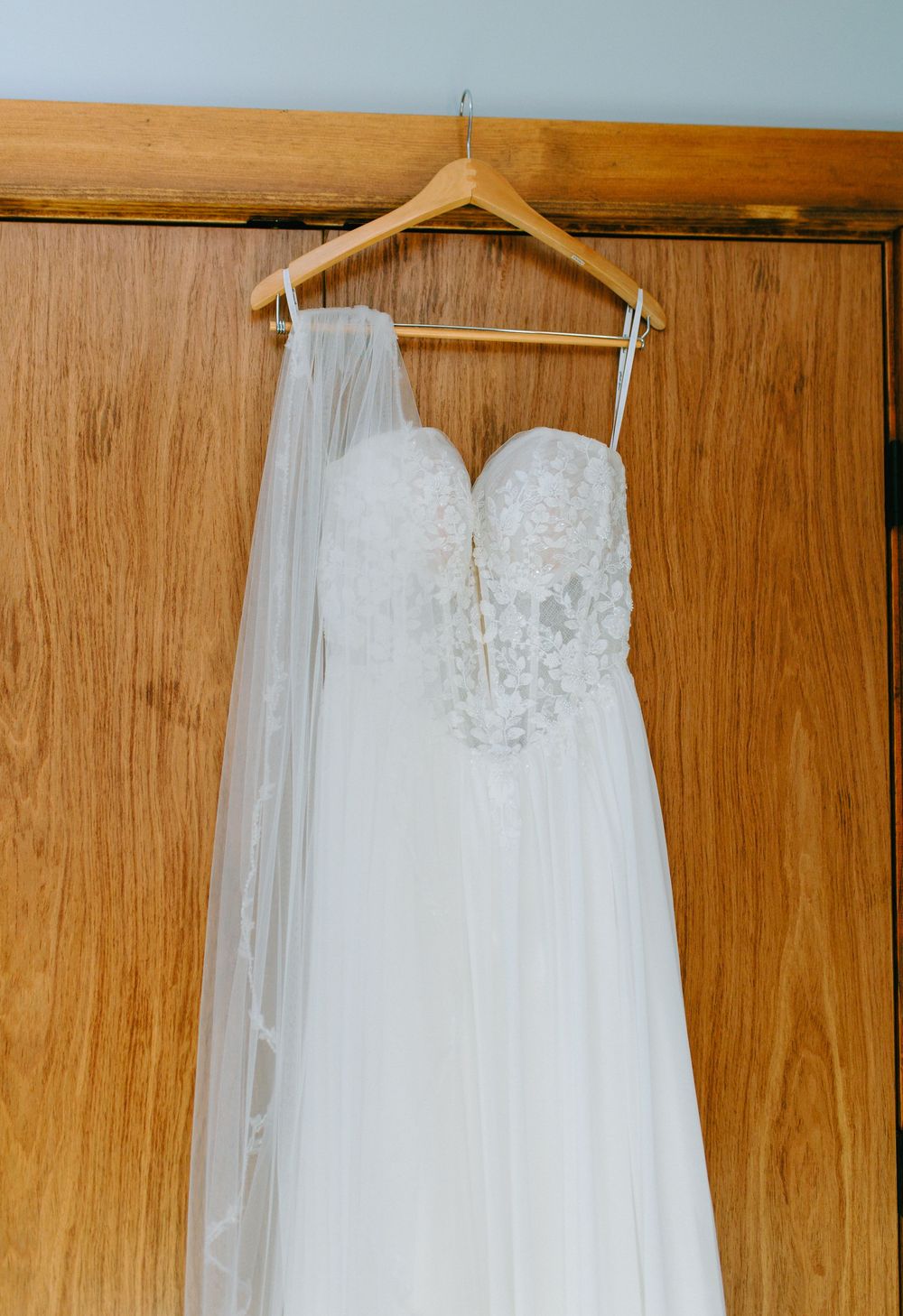 Elegant white wedding dress with delicate lace details hangs on wooden hangers against a light wood paneled wall.