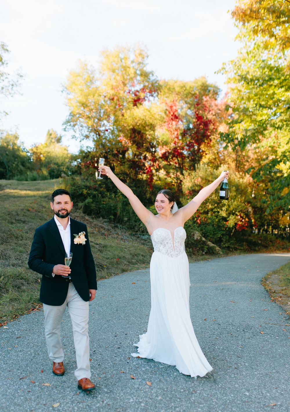 Couple celebrates joyfully on a path surrounded by vibrant autumn foliage during their wedding photoshoot.