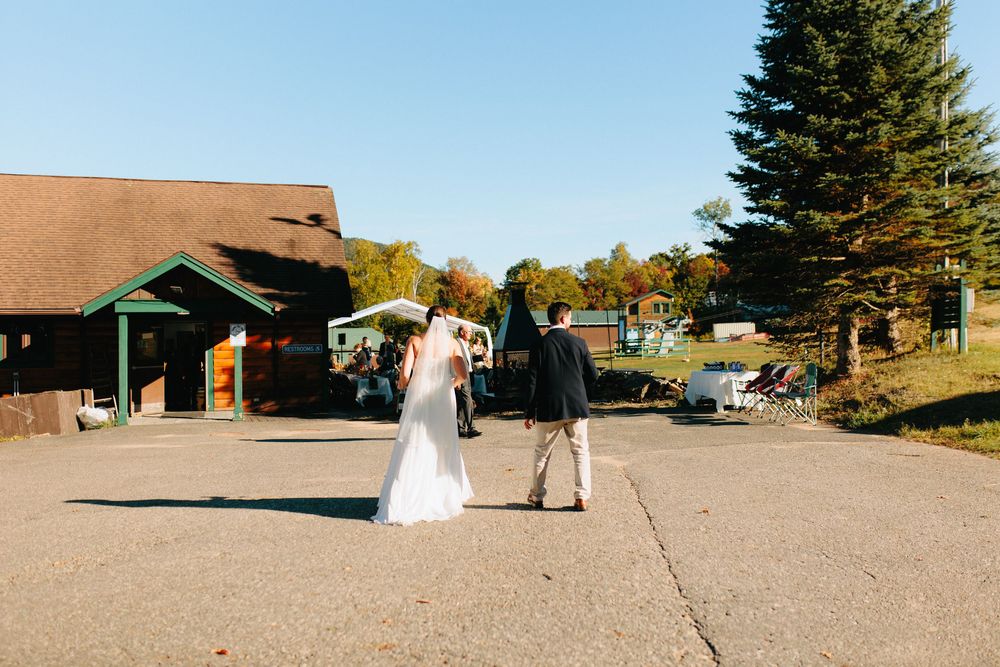 A couple in formal attire walking together outside a rustic lodge building on a sunny autumn day.