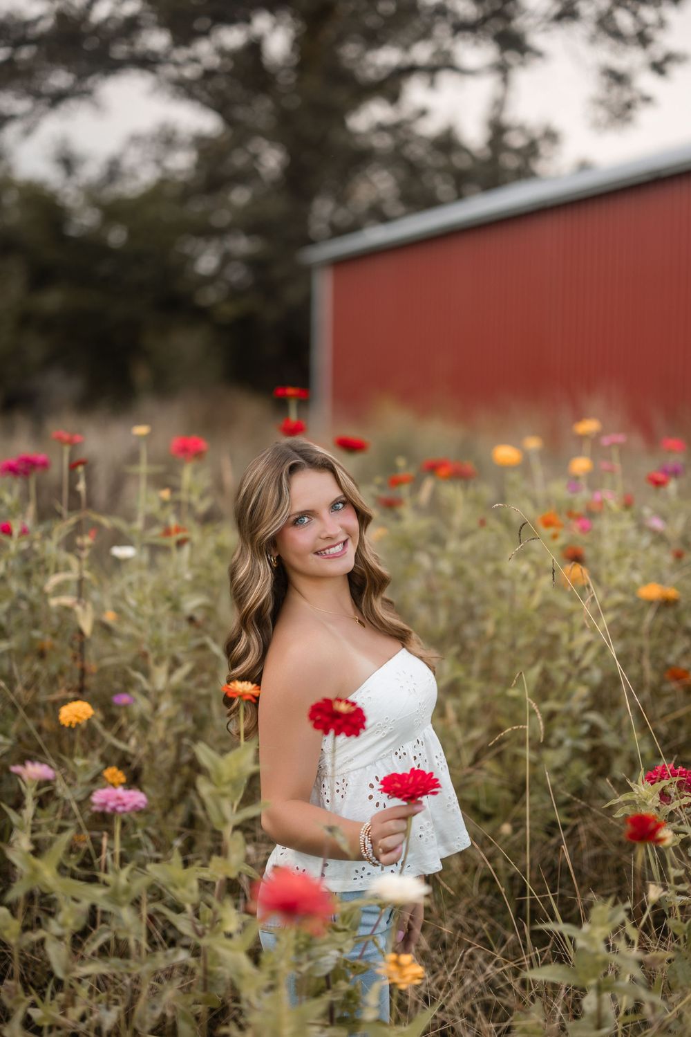 A person holds a red flower while posing in a vibrant zinnia garden with a red barn in the background.