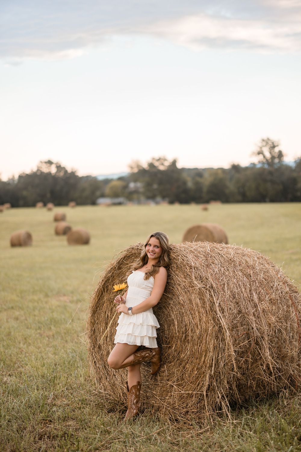 Woman in white dress poses by hay bale in rustic farm field during golden hour sunset.