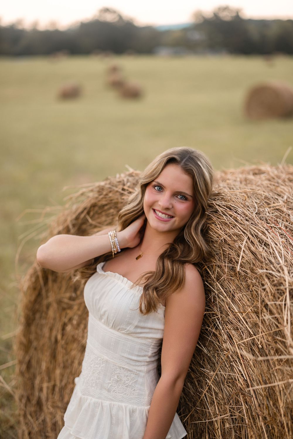 A portrait of someone in a white dress posing against a rustic hay bale at sunset.