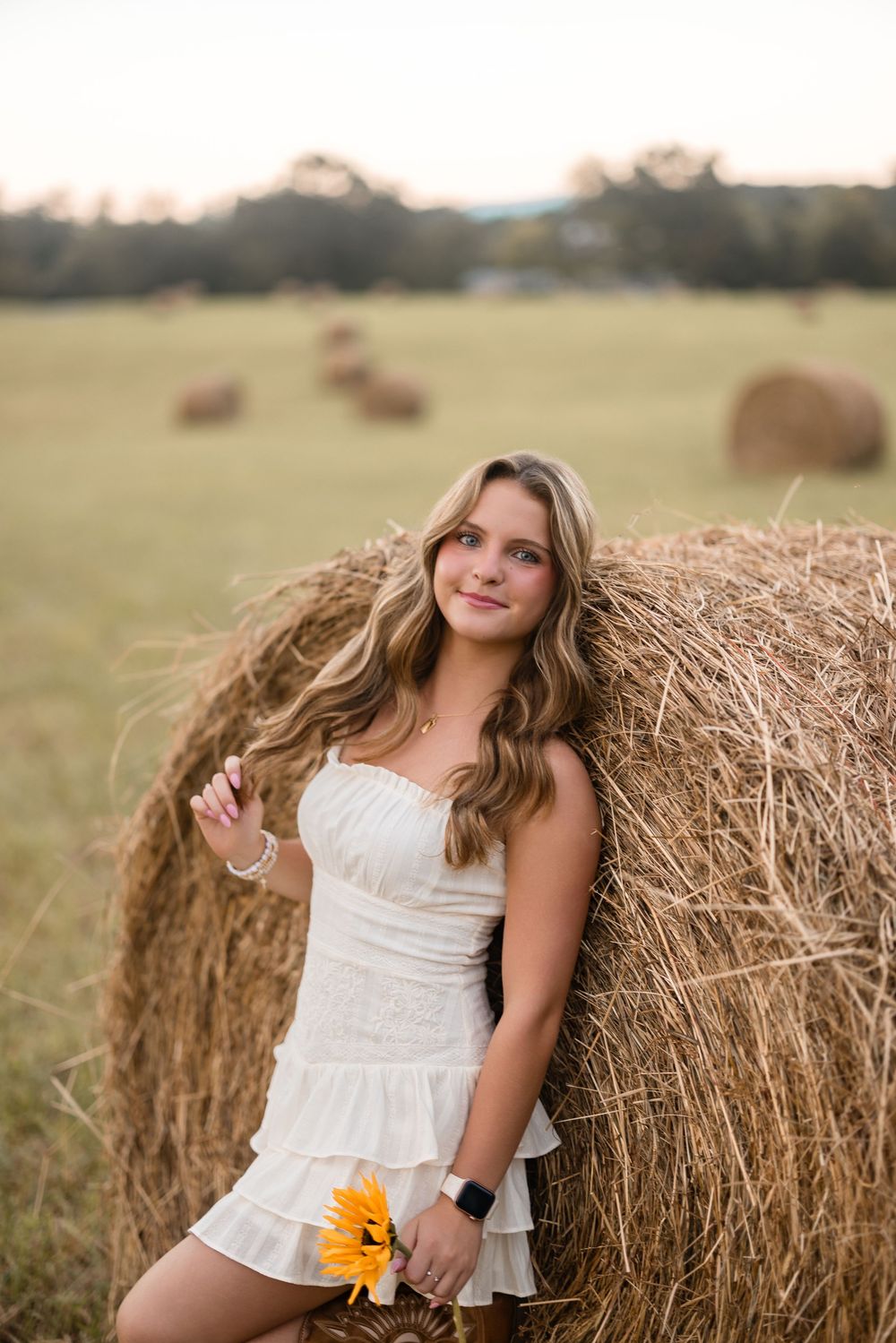 A young girl in a white dress poses near hay bales in a golden field during a portrait photo session.