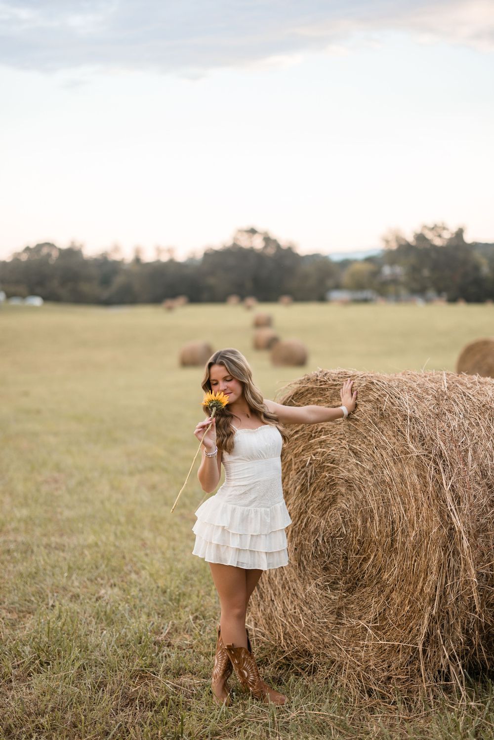 A child in a white dress stands playfully beside a large round hay bale in a rustic farm field setting.