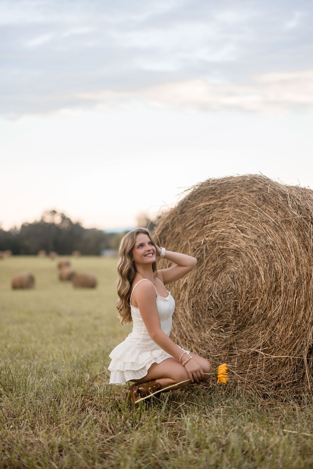 Cheerful person in a white dress sitting and posing on grass near hay bales during golden hour.
