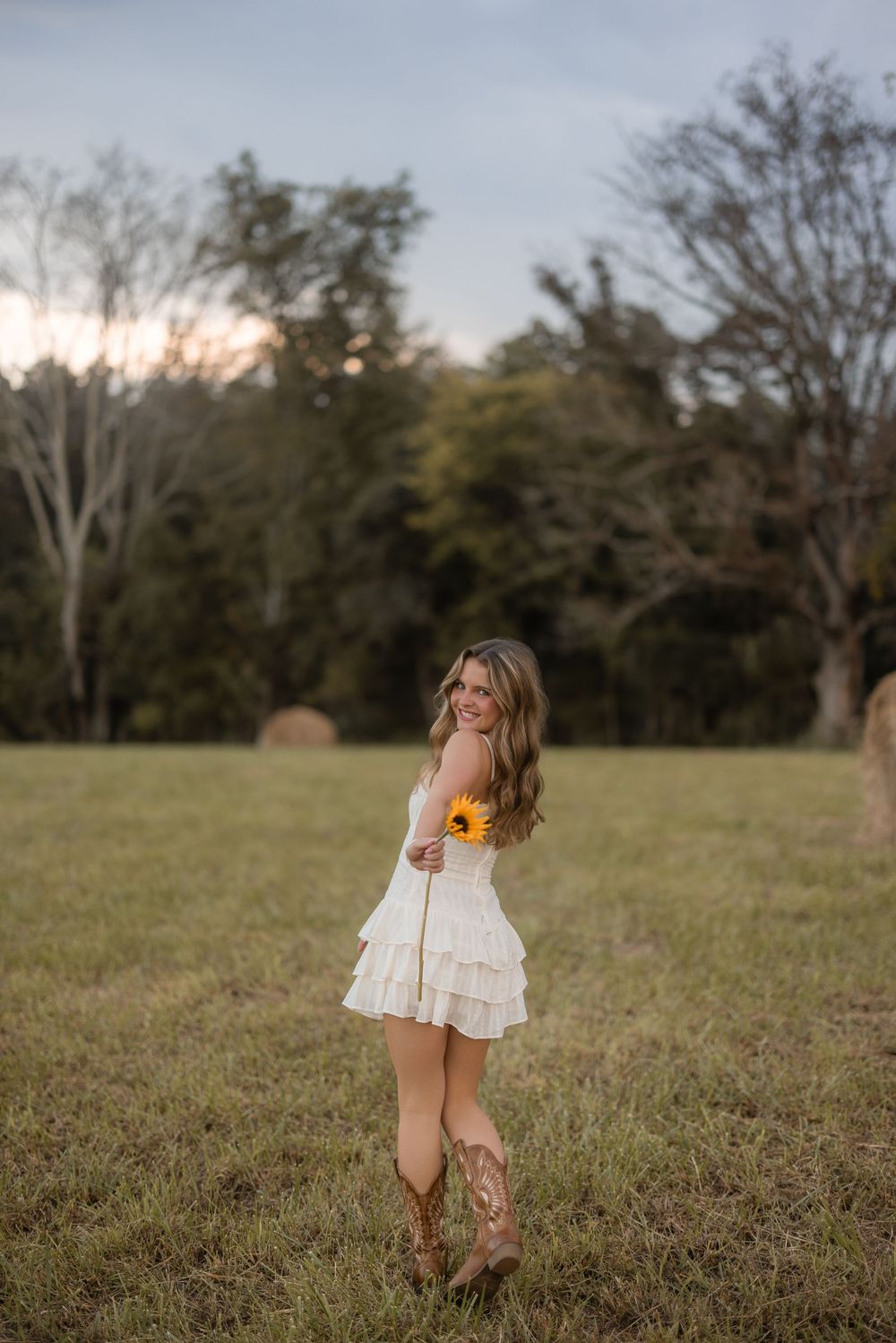 A figure in a white dress stands in a countryside field at dusk holding sunflowers.