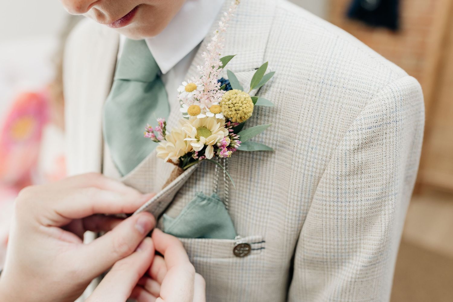 Delicate floral boutonniere being pinned to a light beige textured suit jacket with sage green tie.