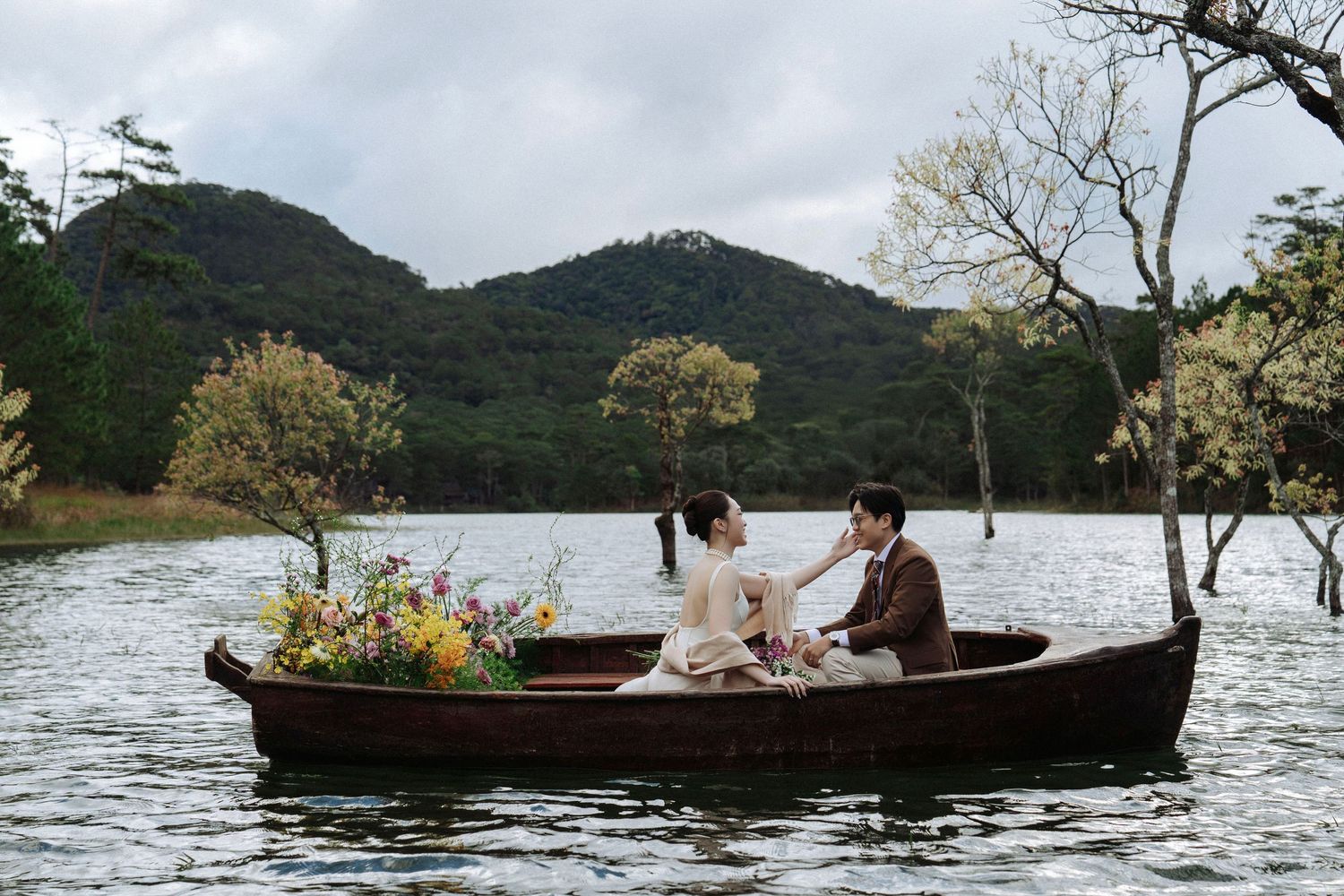 Romantic couple sitting in a wooden boat surrounded by wildflowers on a serene lake with lush hills.