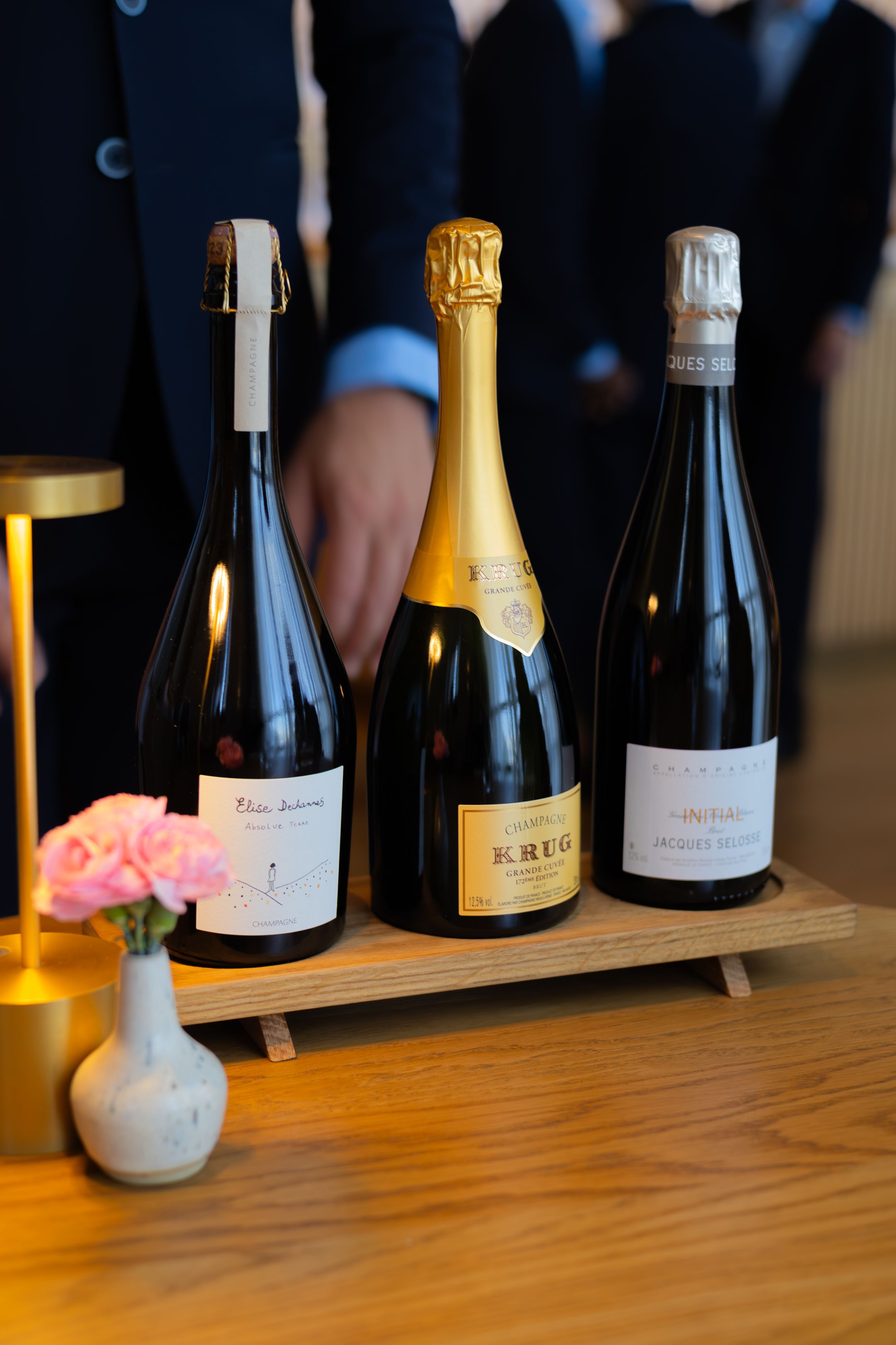 Multiple wine bottles are displayed on a wooden table with a small white vase holding pink flowers in the foreground.