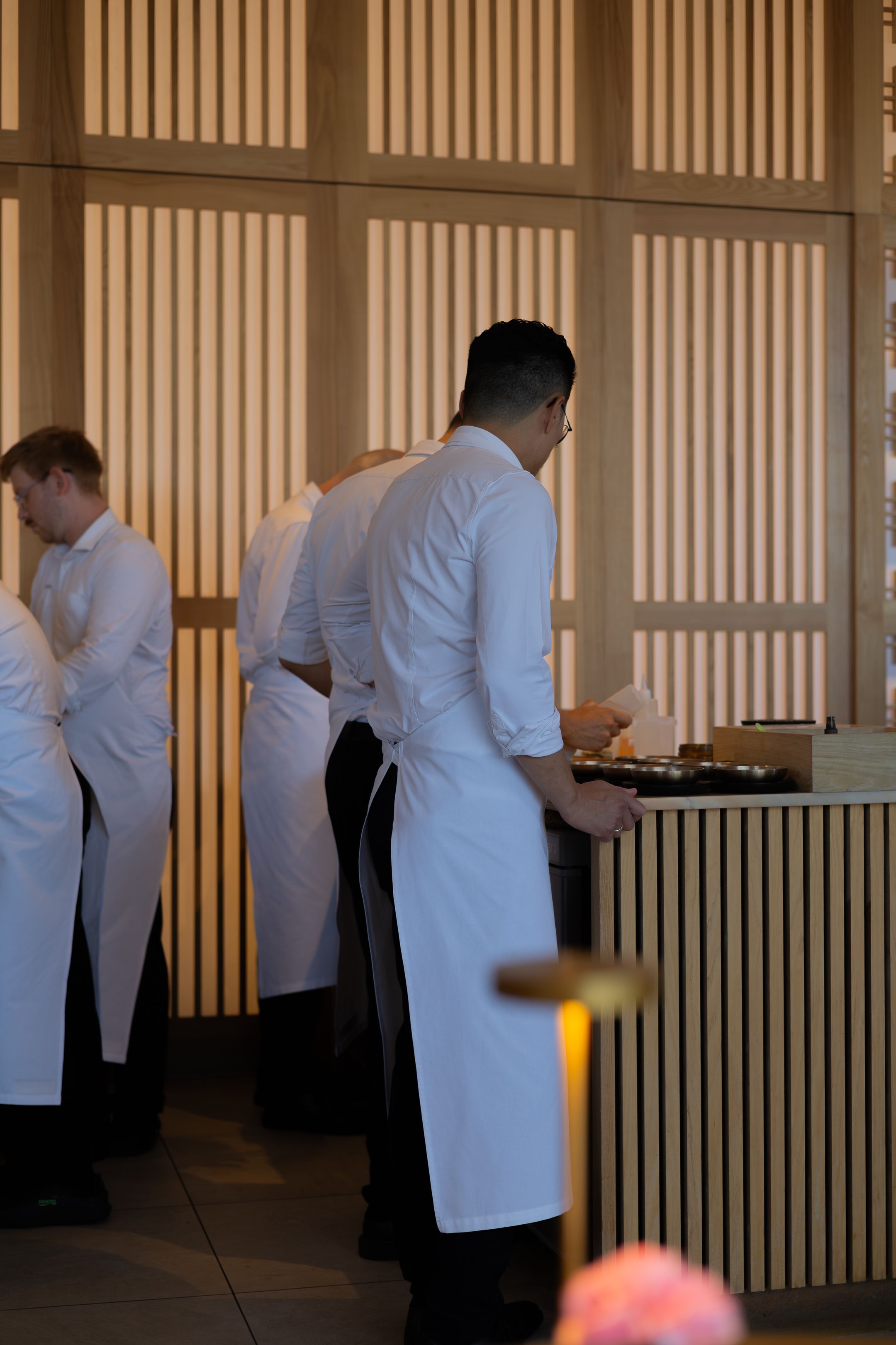 Chefs in white coats working in an upscale restaurant kitchen with wooden slat wall dividers.