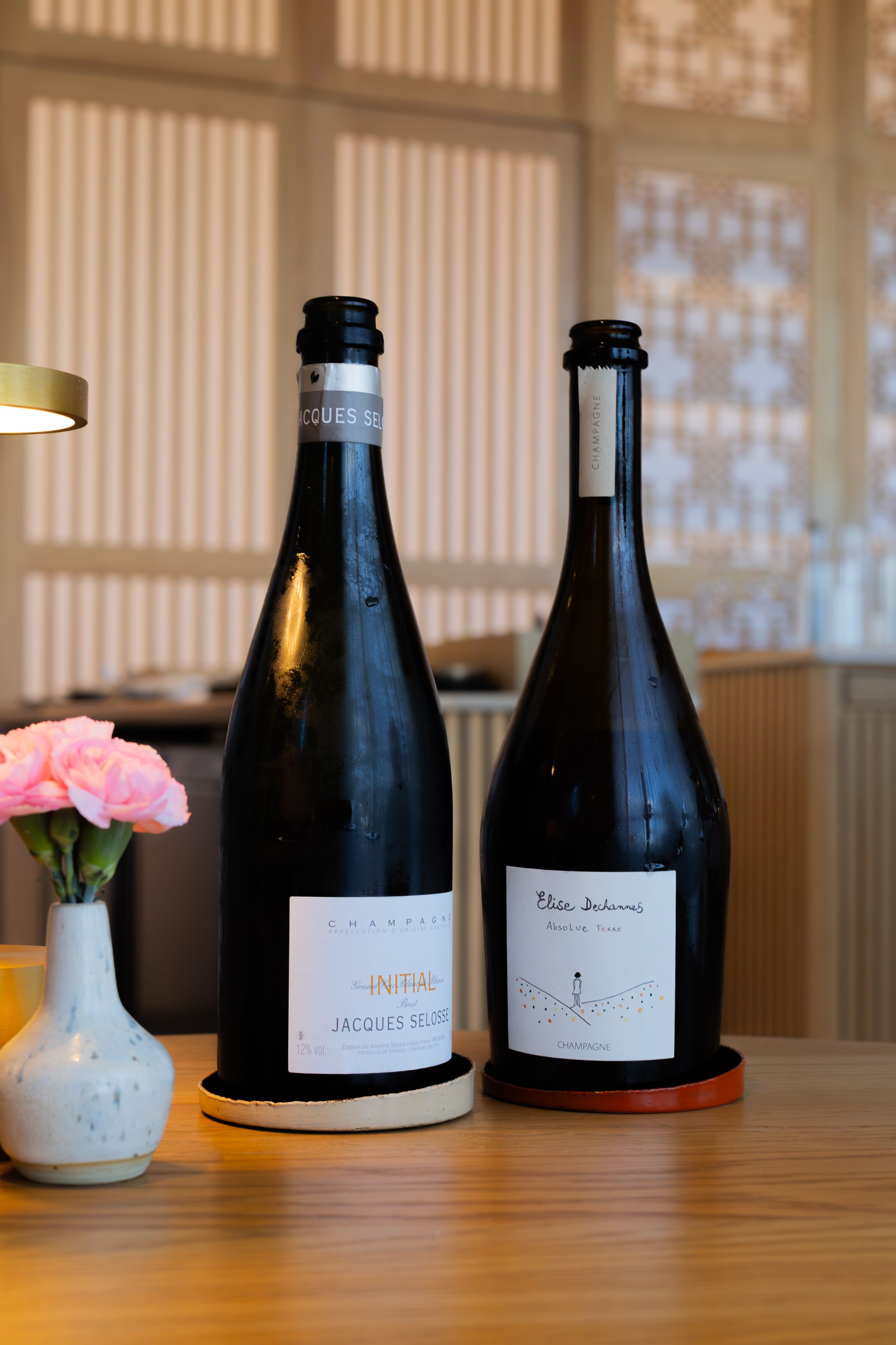 Collection of wine bottles displayed on wooden table with pink flowers in vase and modern Japanese decor.