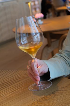 A hand holds a wine glass containing amber-colored wine while examining it against a wooden table surface.