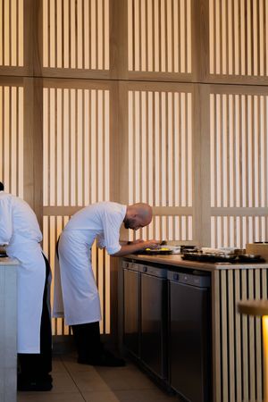 Chef in white coat working at elegant restaurant counter with wooden slat wall decor.