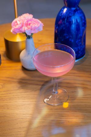 Pink cocktail in a coupe glass alongside blue bottles and candlelight on a wooden surface.