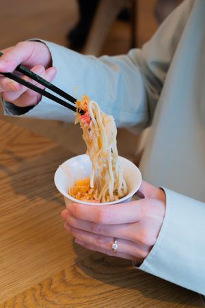Chopsticks lifting steaming noodles from a small white bowl on wooden table.