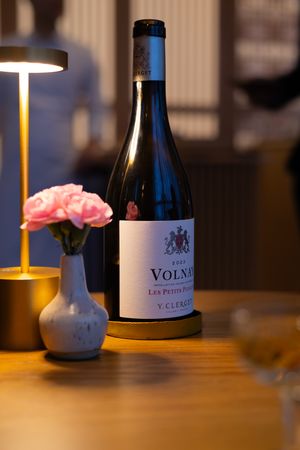 Wine bottles and a pink flower vase illuminated by warm lighting on a wooden table.