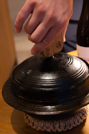 Hands lifting black decorative lid off dish on wooden dining table.