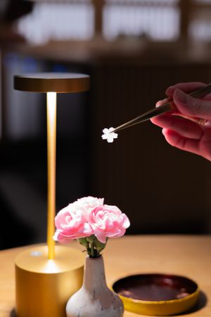 Chopsticks being used over a bowl with pink flowers and wine bottles in background.