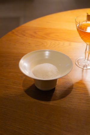 White curved bowl on wooden table with wine glass in soft lighting.