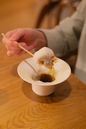 Hands using chopsticks to eat from a white ceramic bowl on wooden table.
