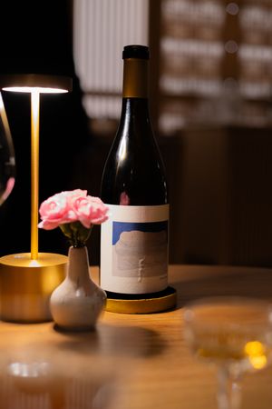 Wine bottle with minimalist label next to small vase and gold lamp on wooden table.