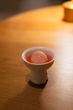 A small white ceramic sake cup containing a pink-colored beverage on a wooden table surface.