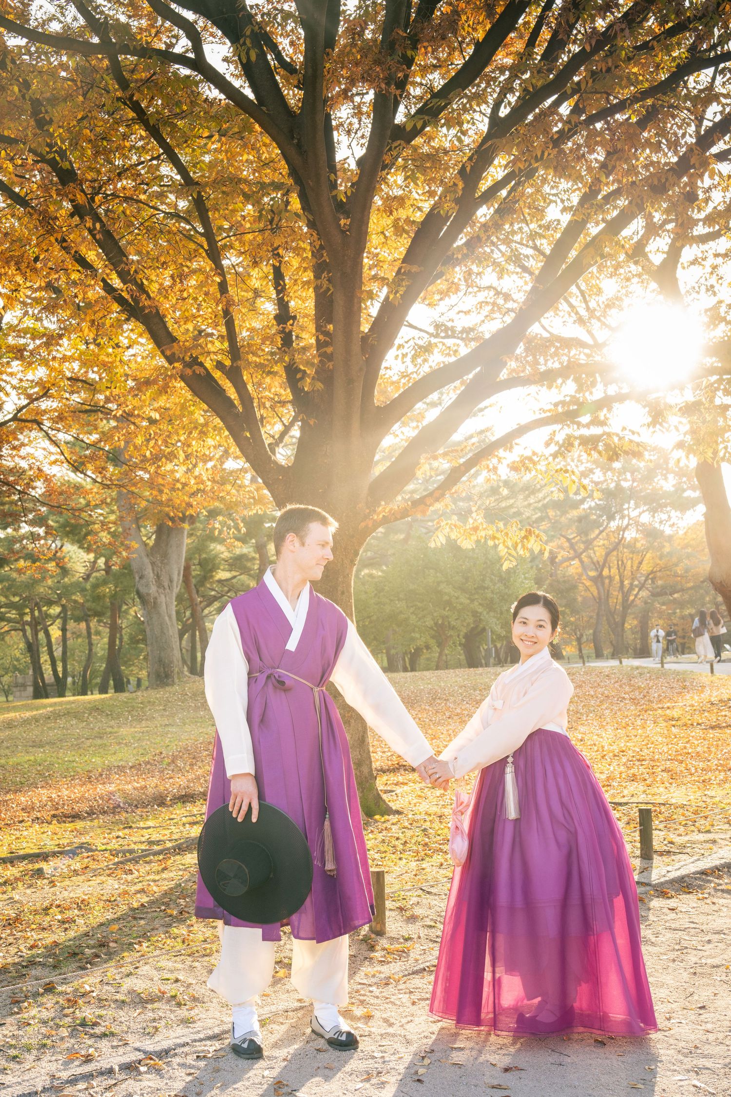People in traditional Korean hanbok attire stand beneath golden autumn trees during sunset in a park.