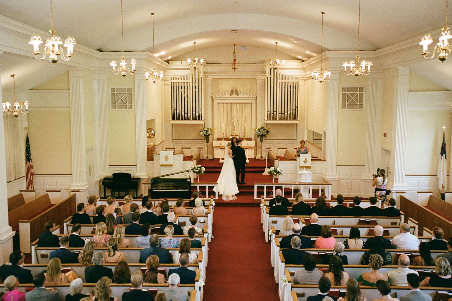 Interior panorama of a traditional church sanctuary with white walls, wooden pews, and chandeliers during a service.