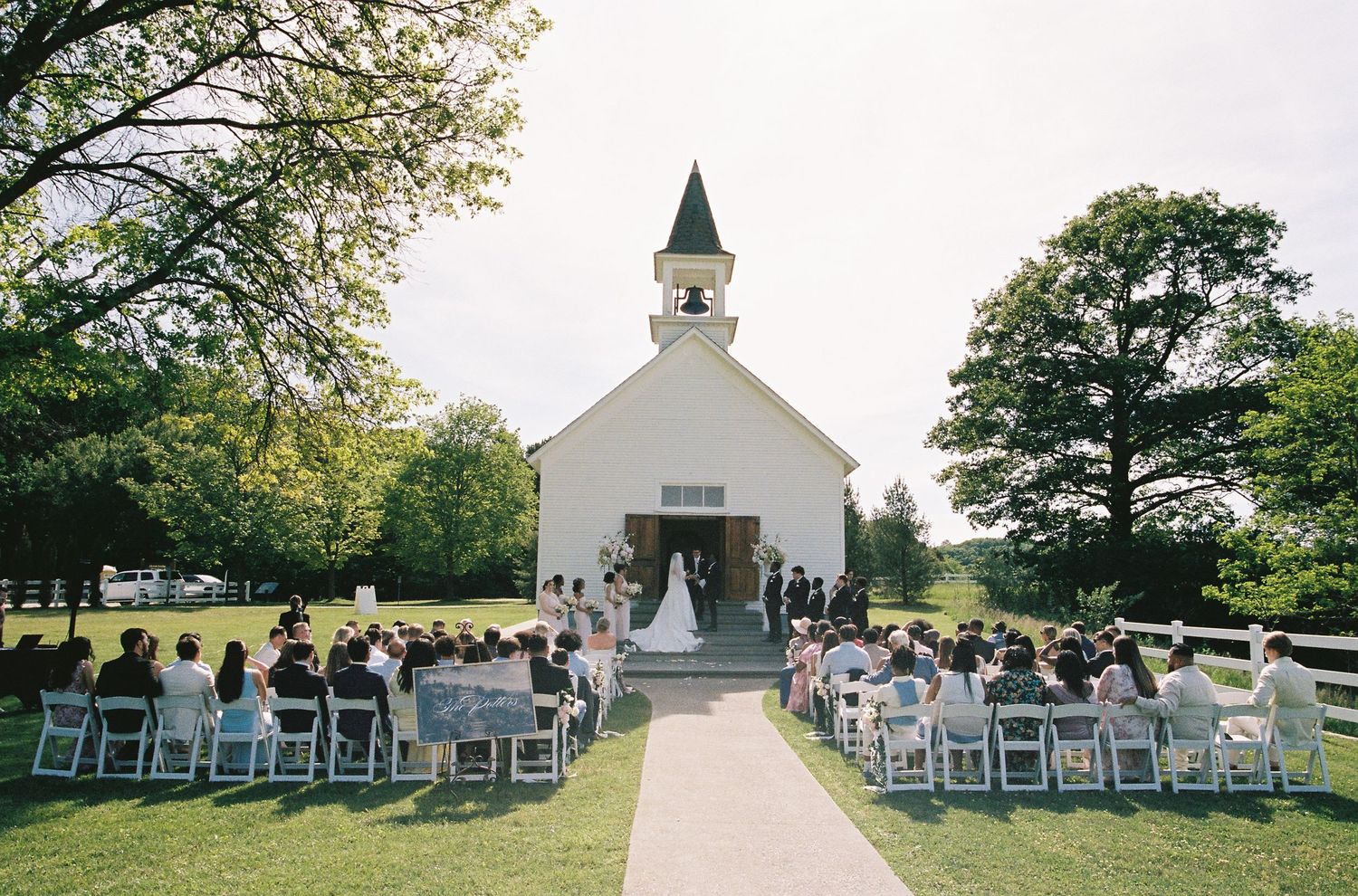 Outdoor wedding ceremony at a white country chapel with guests seated on white chairs along a tree-lined aisle.