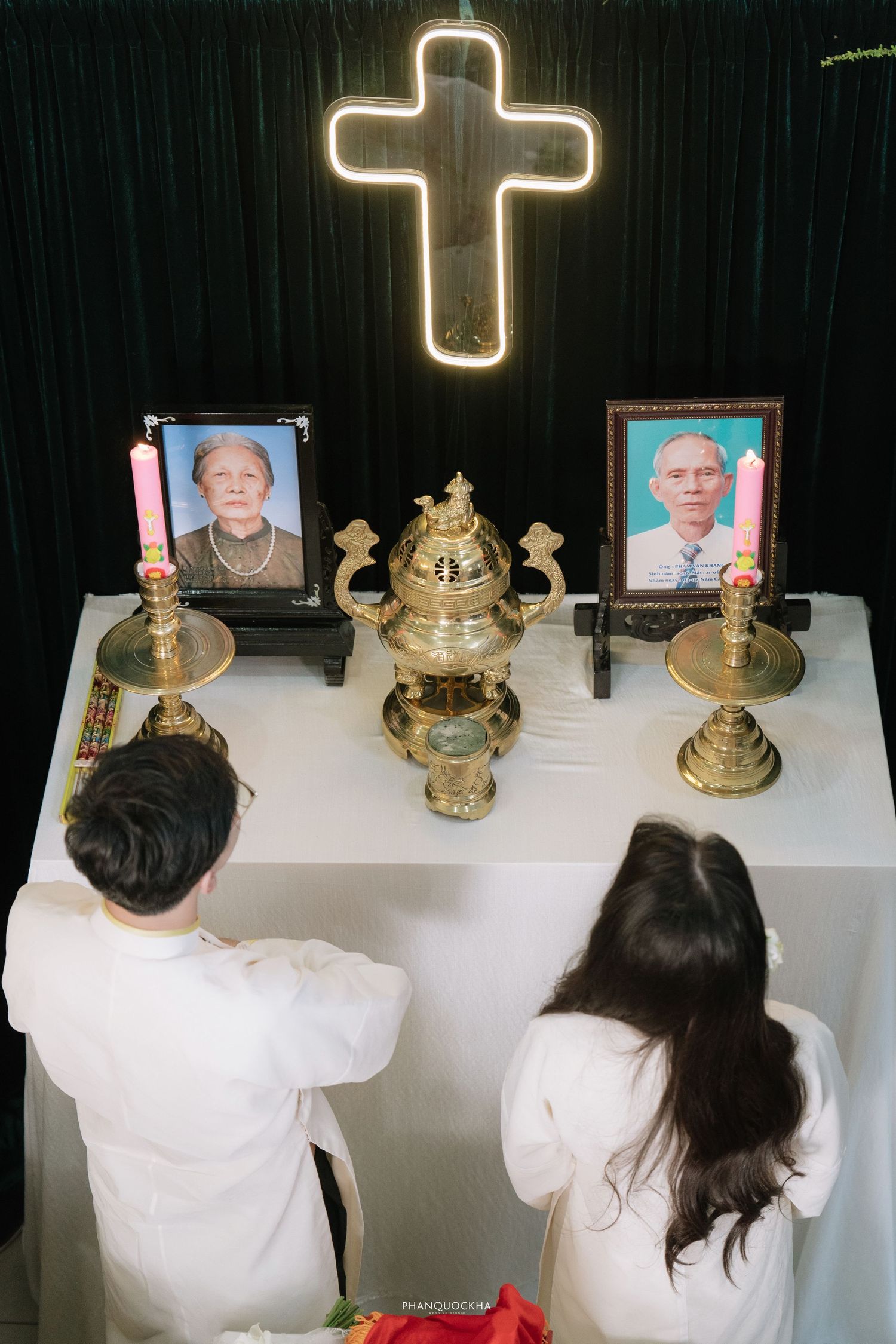 Couple kneels before a glowing cross altar with portraits and candles at a memorial ceremony.