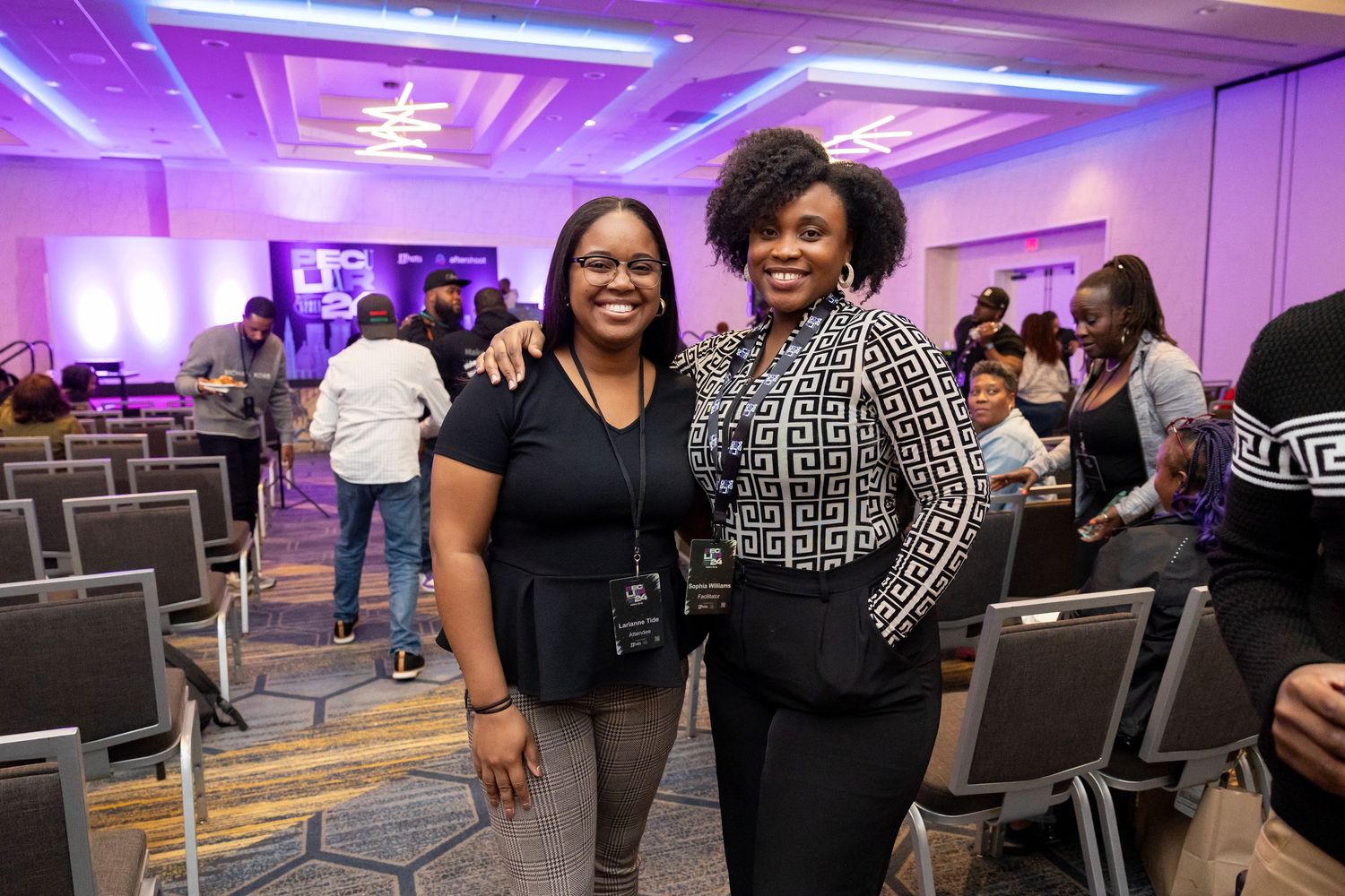 Two smiling women posing together in a purple-lit conference ballroom with attendees in the background.