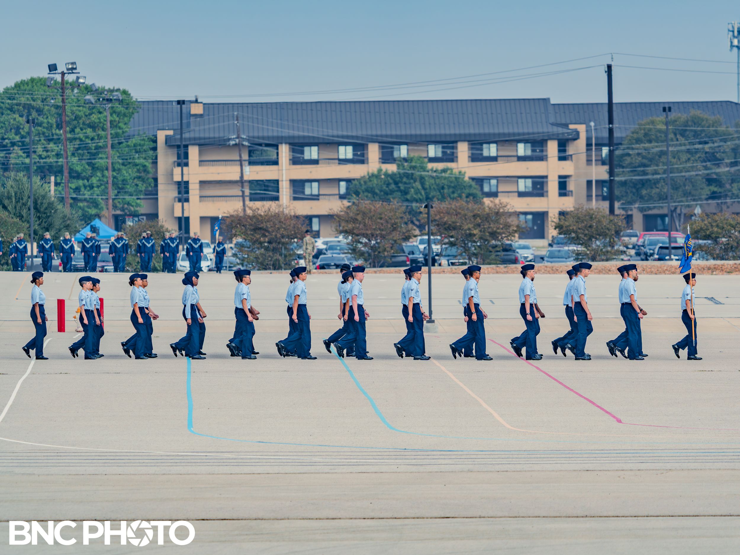Row of U.S. Air Force trainees in light blue shirts marching in single file formation on the parade field during basic training.