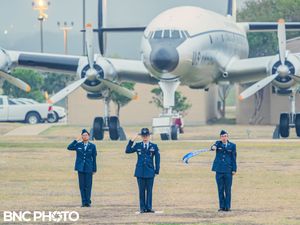 Three U.S. Air Force members in blue dress uniforms saluting on the parade field in front of a vintage aircraft during a graduation ceremony