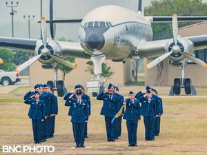 Three U.S. Air Force members in blue dress uniforms saluting on the parade field in front of a vintage aircraft during a graduation ceremony