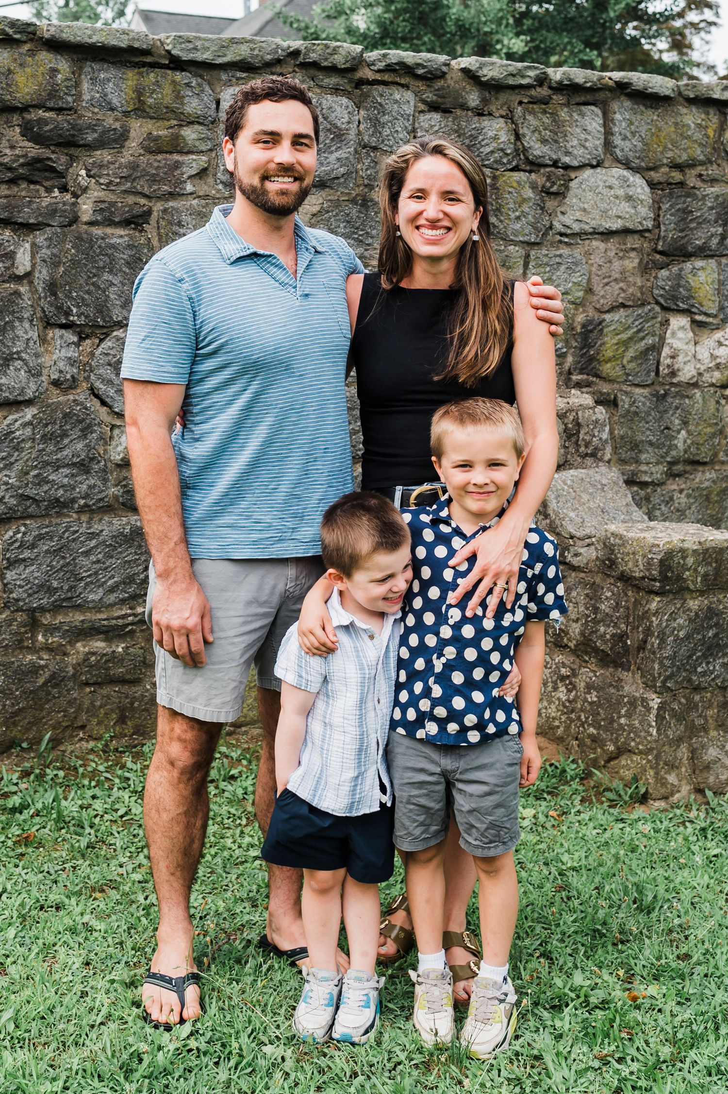 A small group stands together in front of a stone wall on grass, wearing casual summer attire.