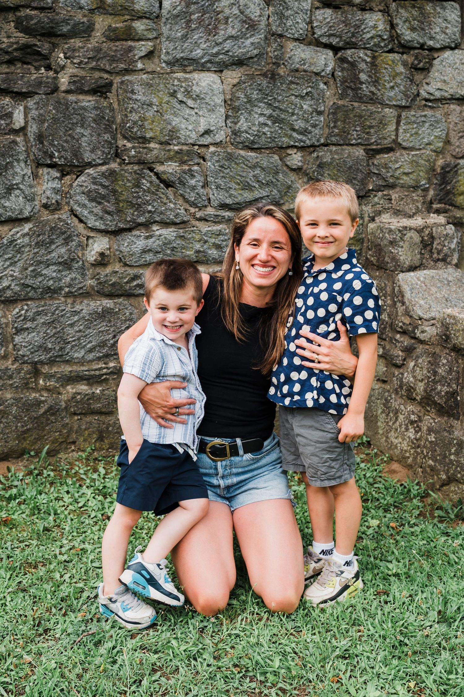 A family sits together on grass against a rustic stone wall background.