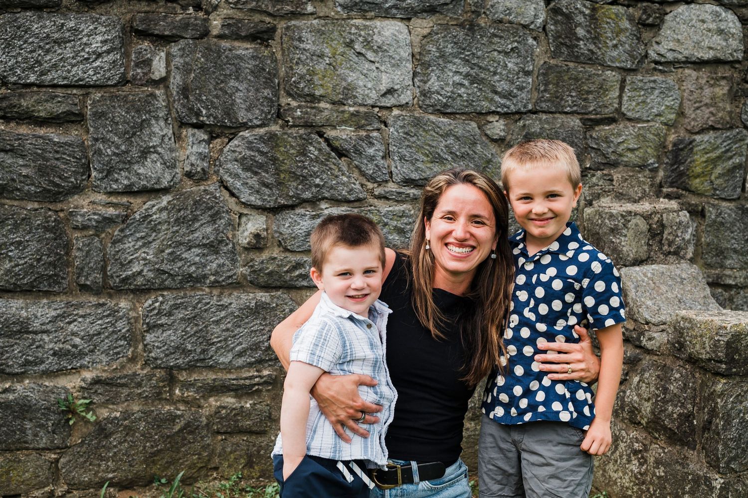 Family laughing and embracing against rustic stone wall background in outdoor portrait session.