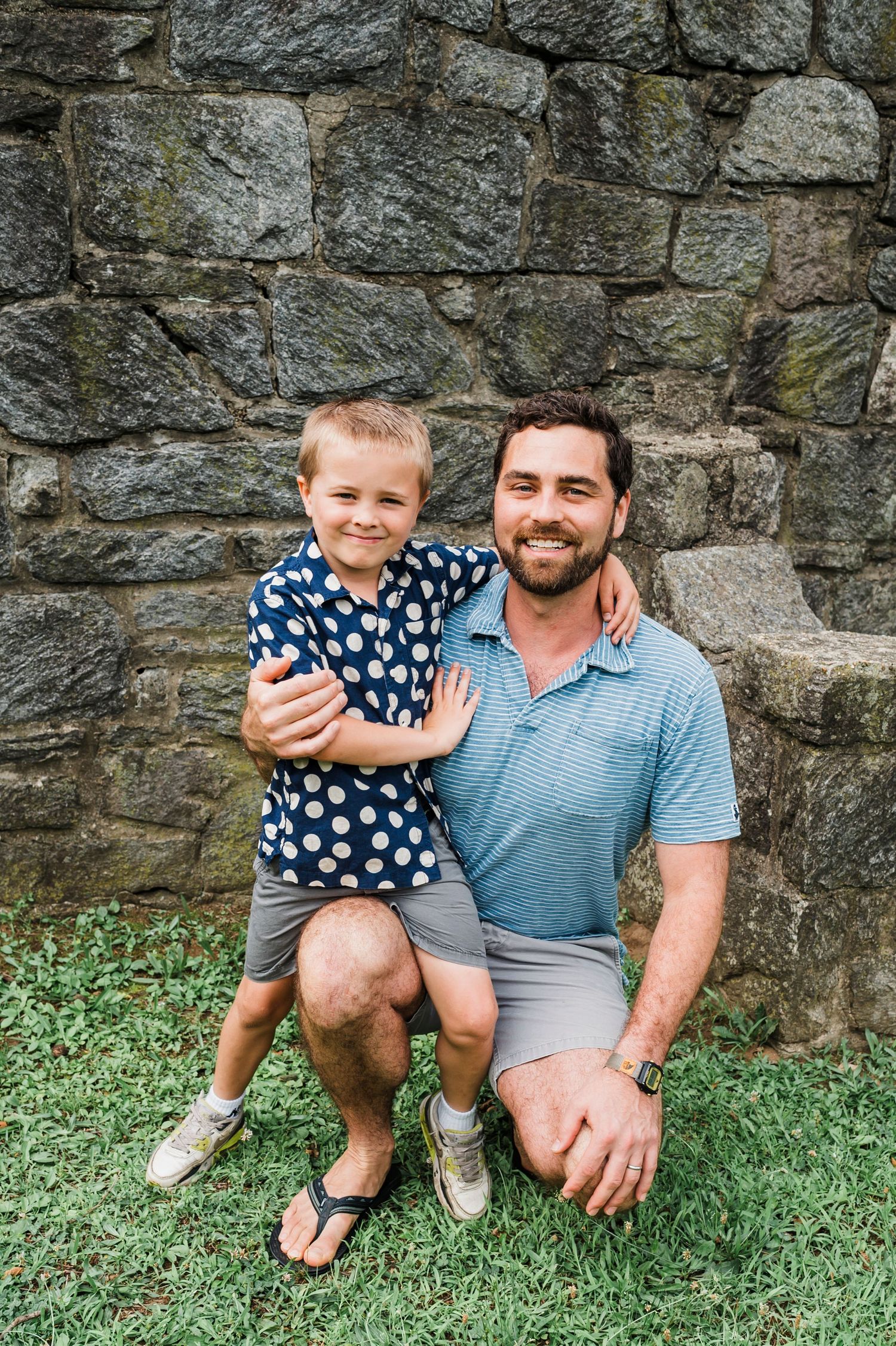 Family enjoying a casual outdoor photo session against a rustic stone wall with green grass.