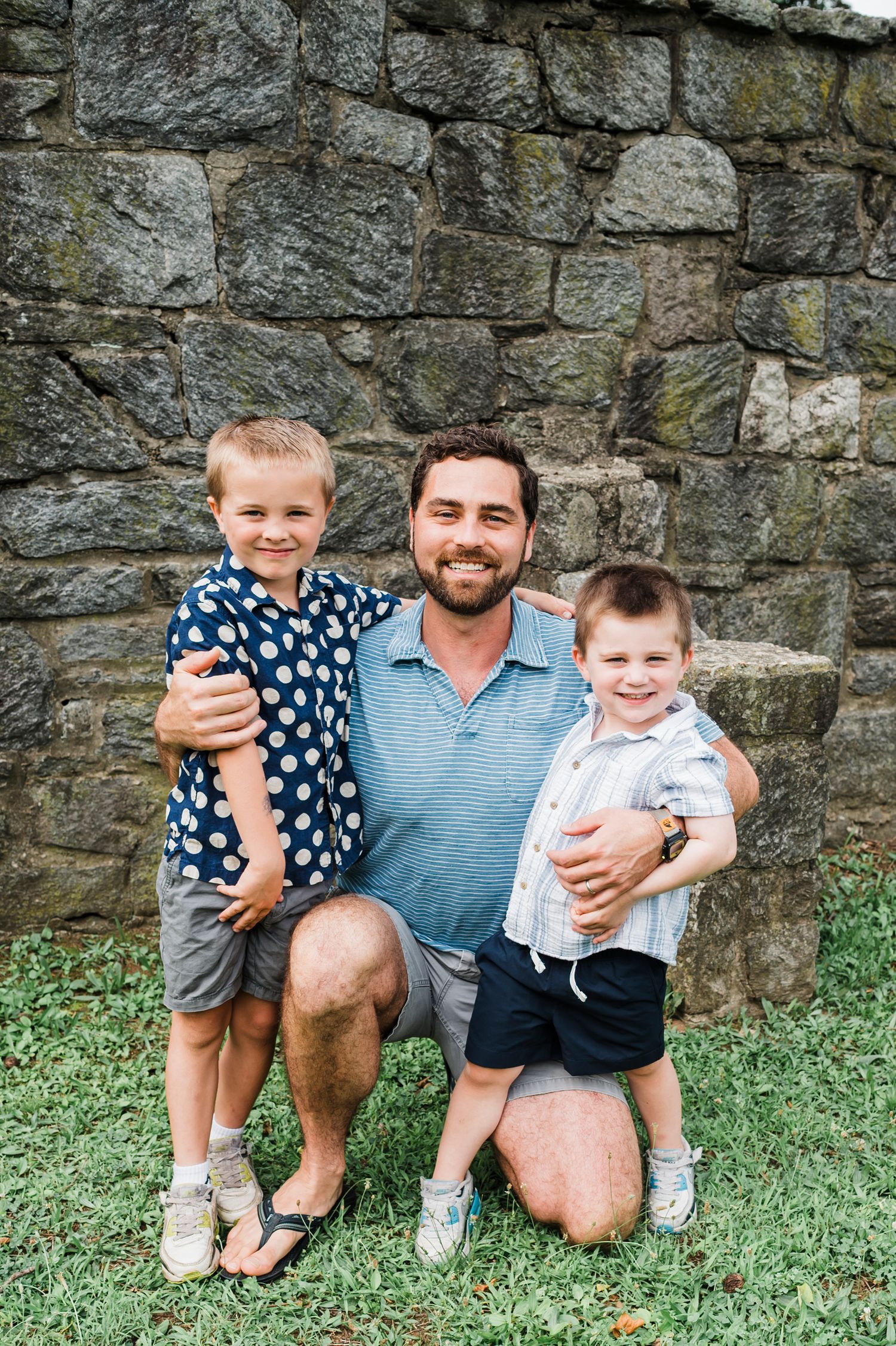 Casual outdoor portrait session with loved ones sitting by stone wall on grass.