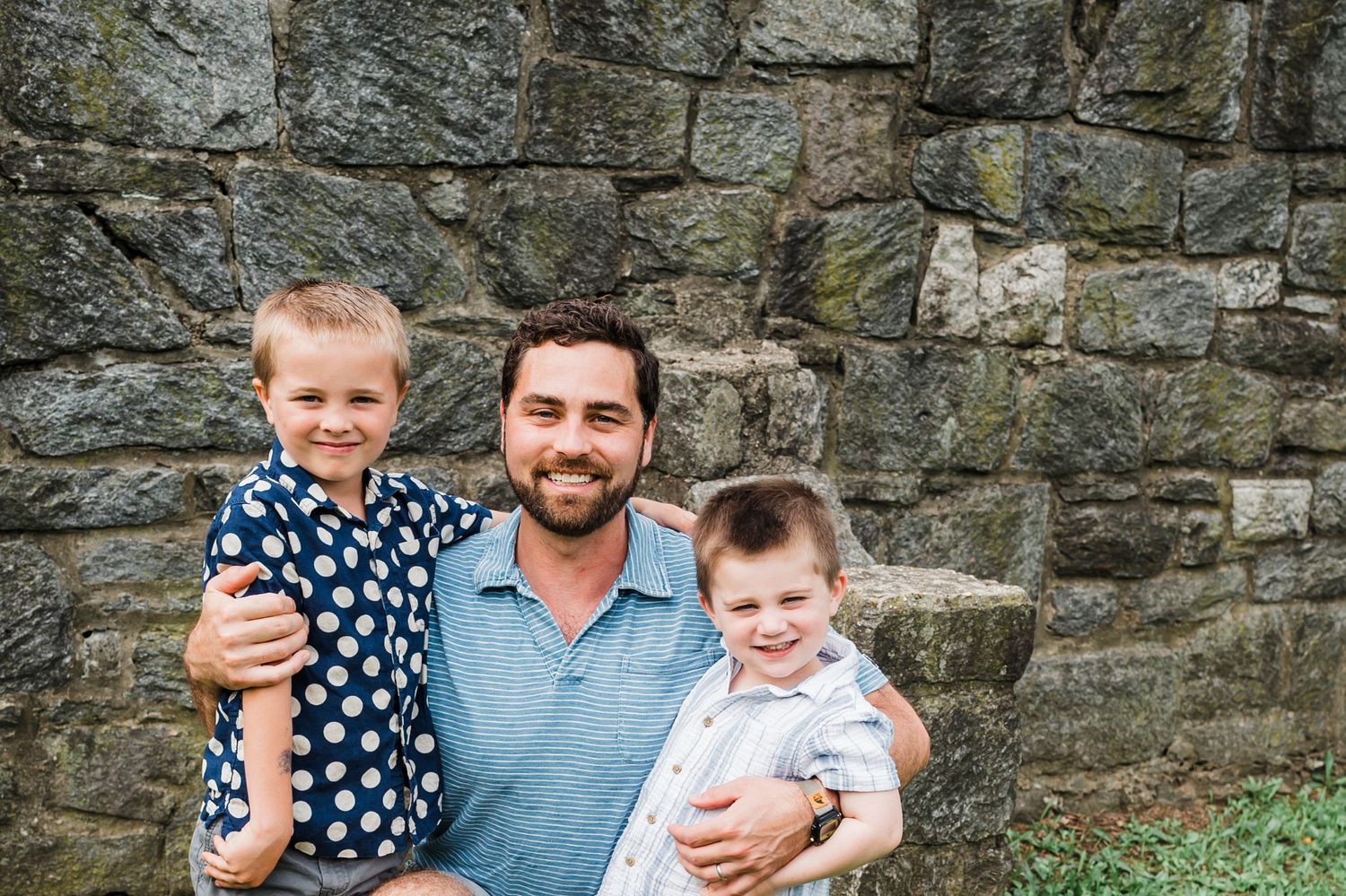 Three people pose together against a rustic stone wall background outdoors.