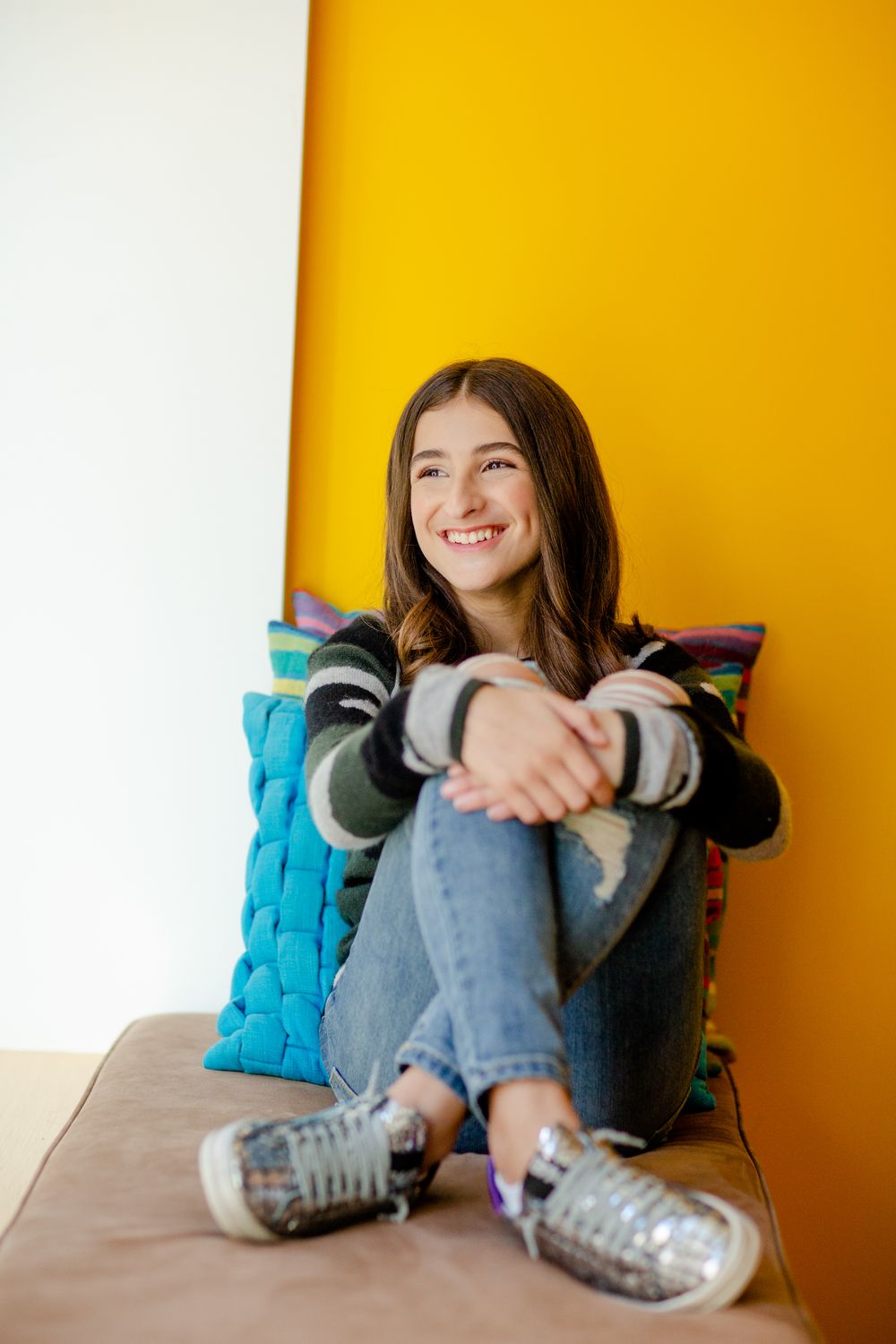 Woman in jeans and sneakers relaxes on a bright yellow couch with turquoise pillows.