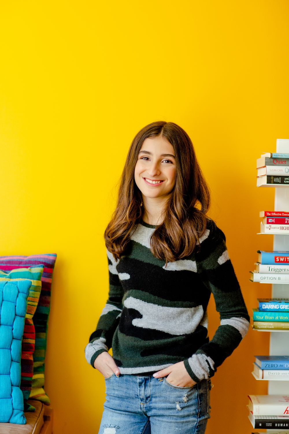 Someone in a striped sweater poses against a bright yellow wall with stacked books.