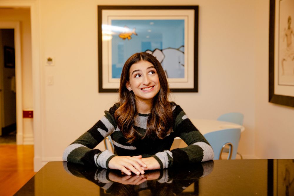 Person smiling while sitting in a casual environment with framed artwork on beige walls.