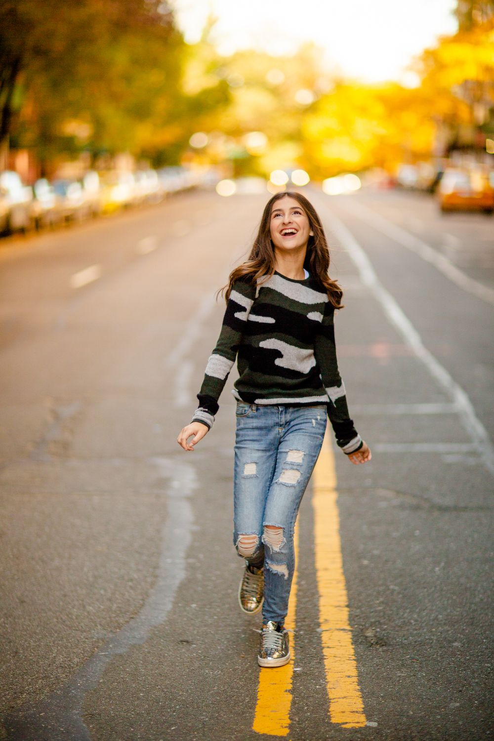 A person in ripped jeans and sweater walks down a street during golden hour sunset.