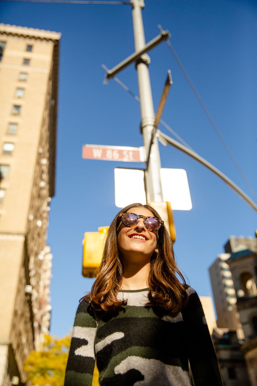 Person smiling in sunlight on urban street corner wearing camouflage sweater.