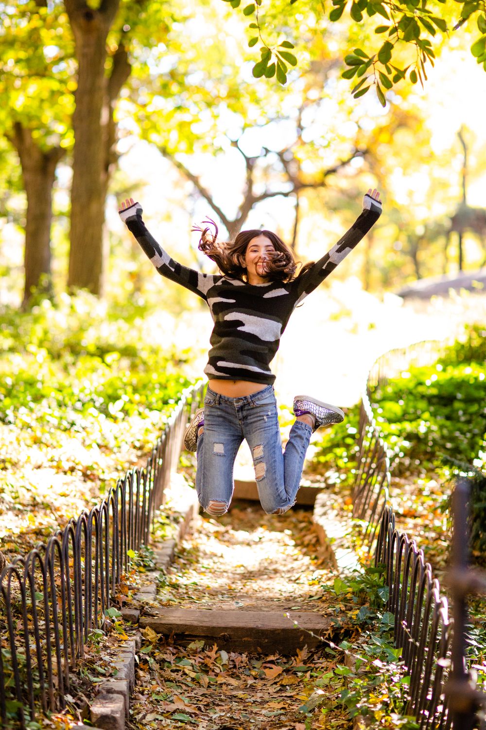 Friends joyfully jumping on a wooded path surrounded by fall foliage and rustic fencing in a park.