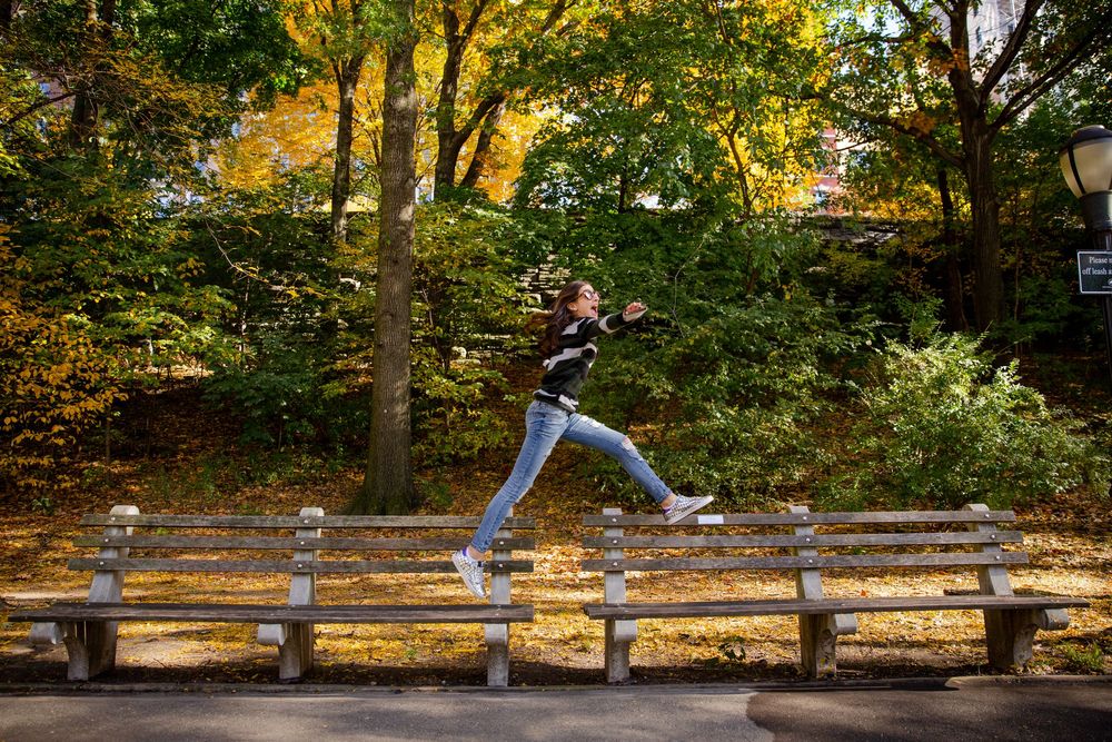 People joyfully dance and leap through a scenic autumn park with golden foliage on trees.