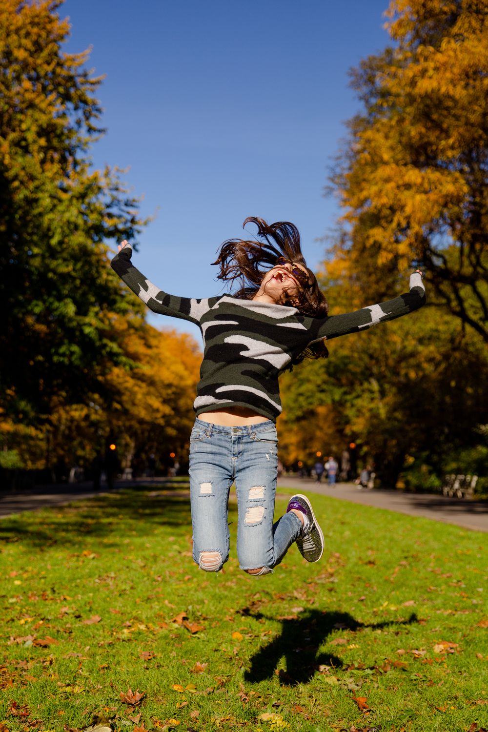 Someone jumps playfully on grass surrounded by autumn trees with golden leaves.